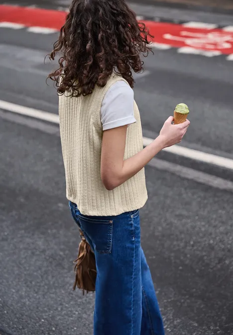 Frau von hinten mit lockigem Haar, trägt weißes T-Shirt, beige Weste, blaue Jeans und hält eine grüne Eistüte.